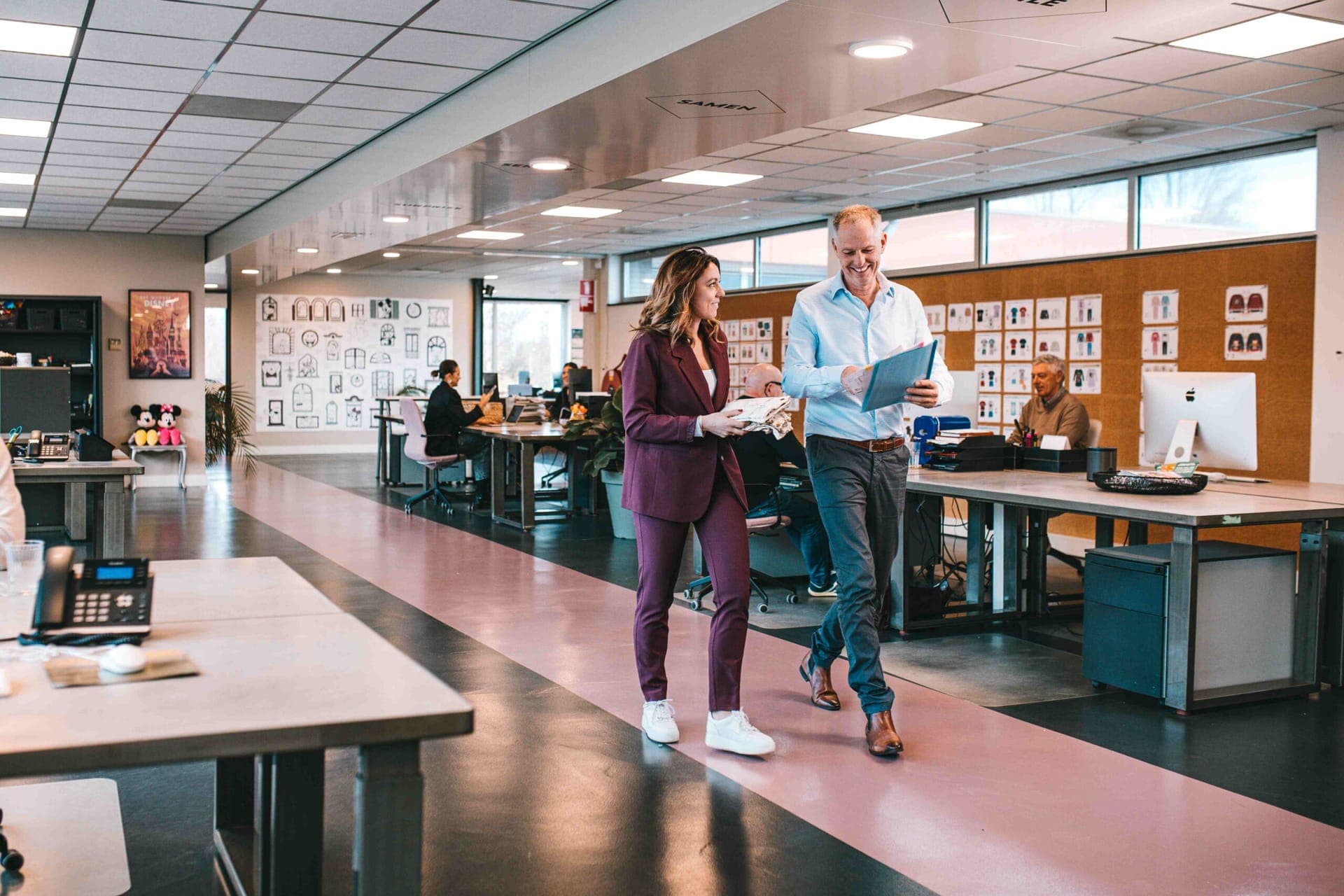 Twee collega's lopen met een map en kledingstalen door het open ontwerpkantoor, met kledingschetsen aan de muur